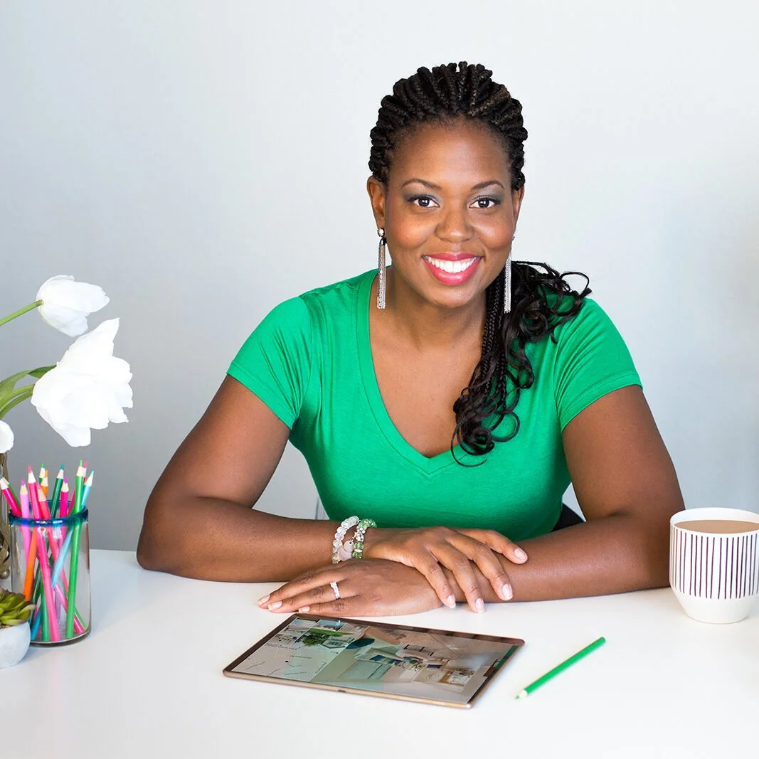 A woman with braided hair, wearing a green shirt, sitting at a white desk with a tablet, colorful pens in a jar, a white flower, a striped mug, and a green pen. She is smiling at the camera.