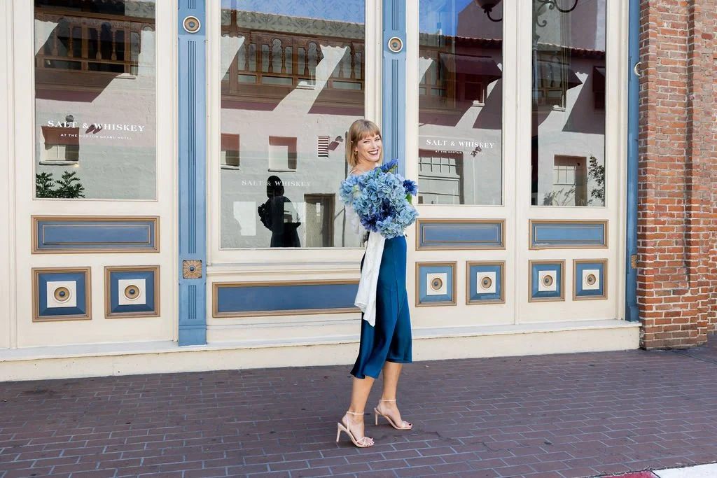 A woman standing on a brick sidewalk in front of a large windowed storefront, holding a large bouquet of blue flowers and smiling, dressed in a dark blue outfit and beige high heels.