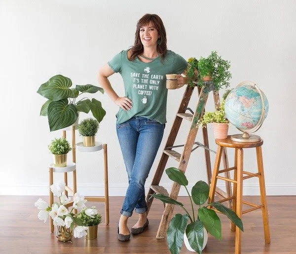 Woman standing in a room decorated with houseplants, a globe, and a wooden ladder near a stool, smiling at the camera.