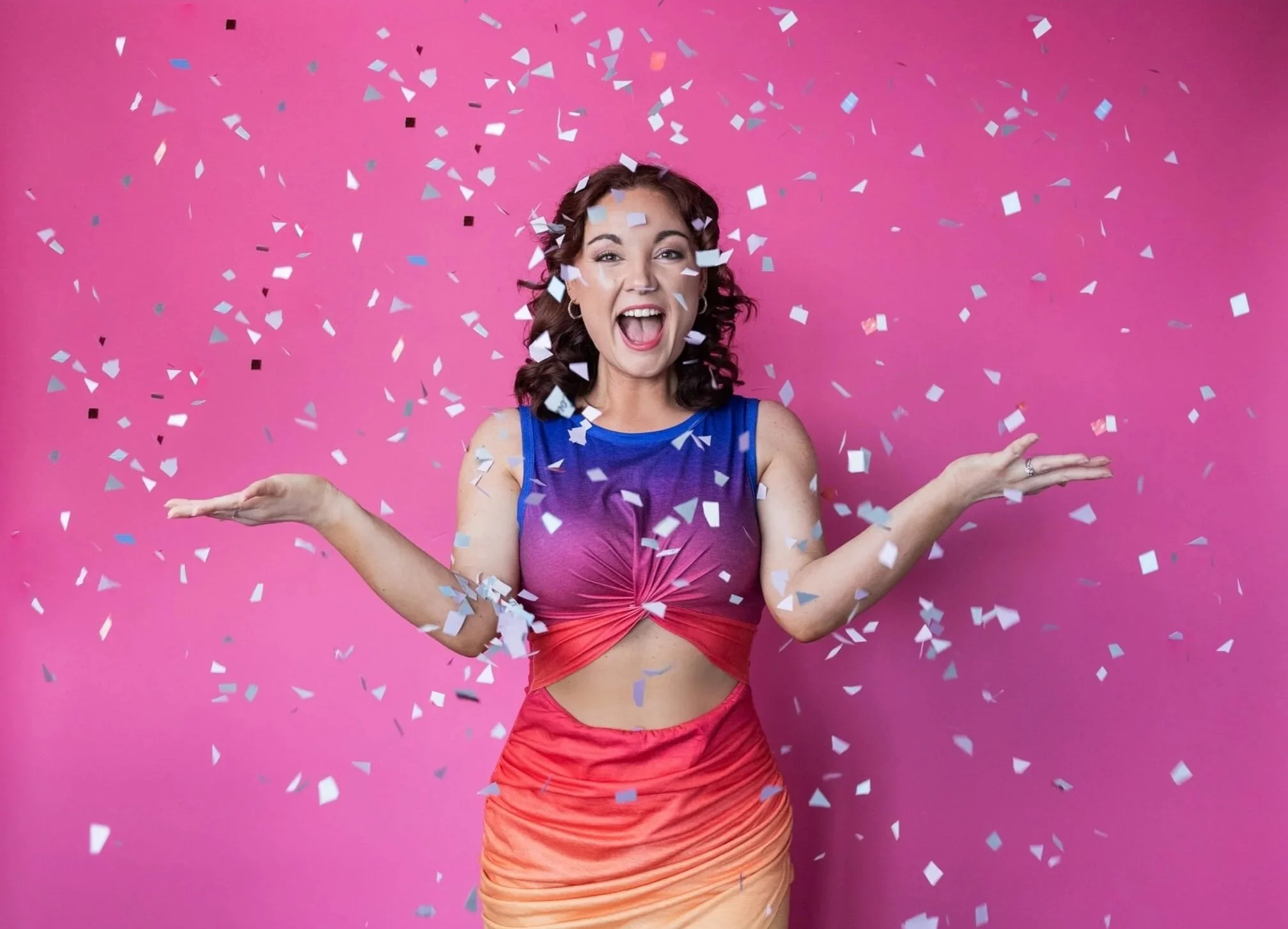 A woman celebrating with hands open, surrounded by showered confetti, against a pink background, wearing a colorful dress.