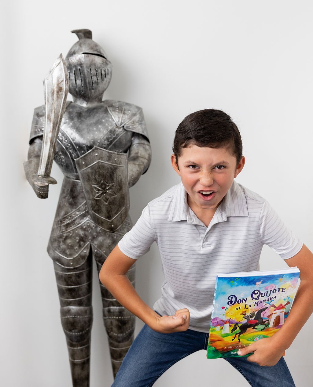 A young boy in a white striped shirt and blue jeans is posing angrily, flexing his right arm and holding a colorful children's book titled 'Don Quijote de la Mancha.' Behind him, there is a large metallic knight statue holding a sword against a plain white wall.