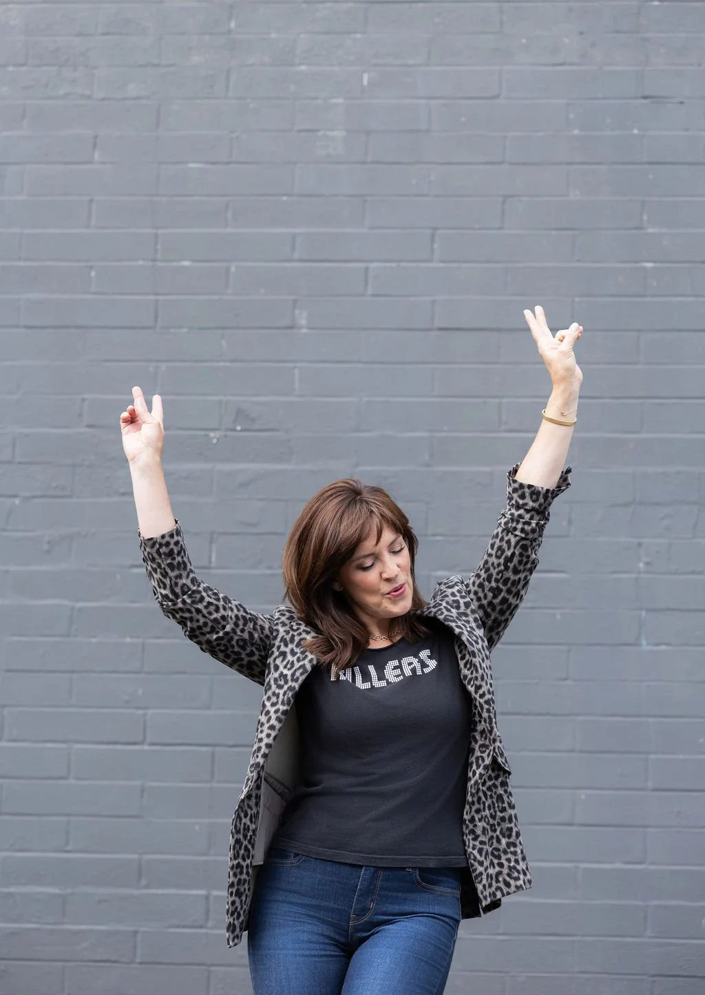 Woman with short brown hair wearing a leopard print blazer and a black T-shirt with the word 'killers' and blue jeans, standing in front of a gray brick wall, with arms raised and eyes closed.