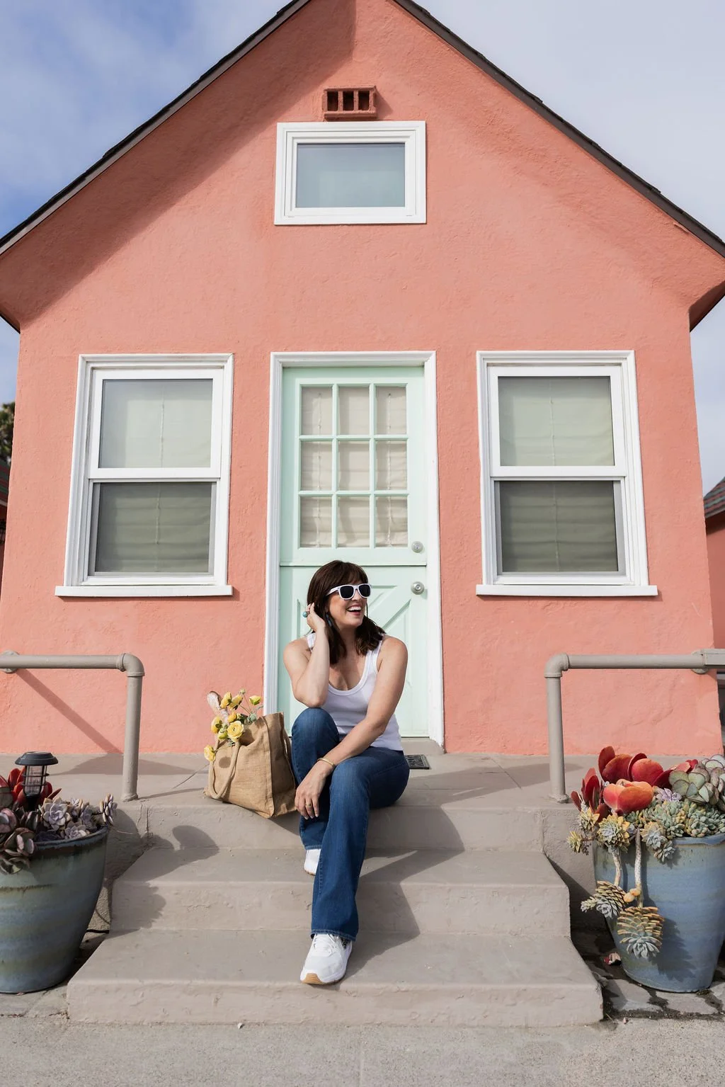 A woman sitting on the steps of a pink house with white-framed windows and a light green door, wearing sunglasses, a white tank top, and blue jeans, with a bag of flowers at her side.
