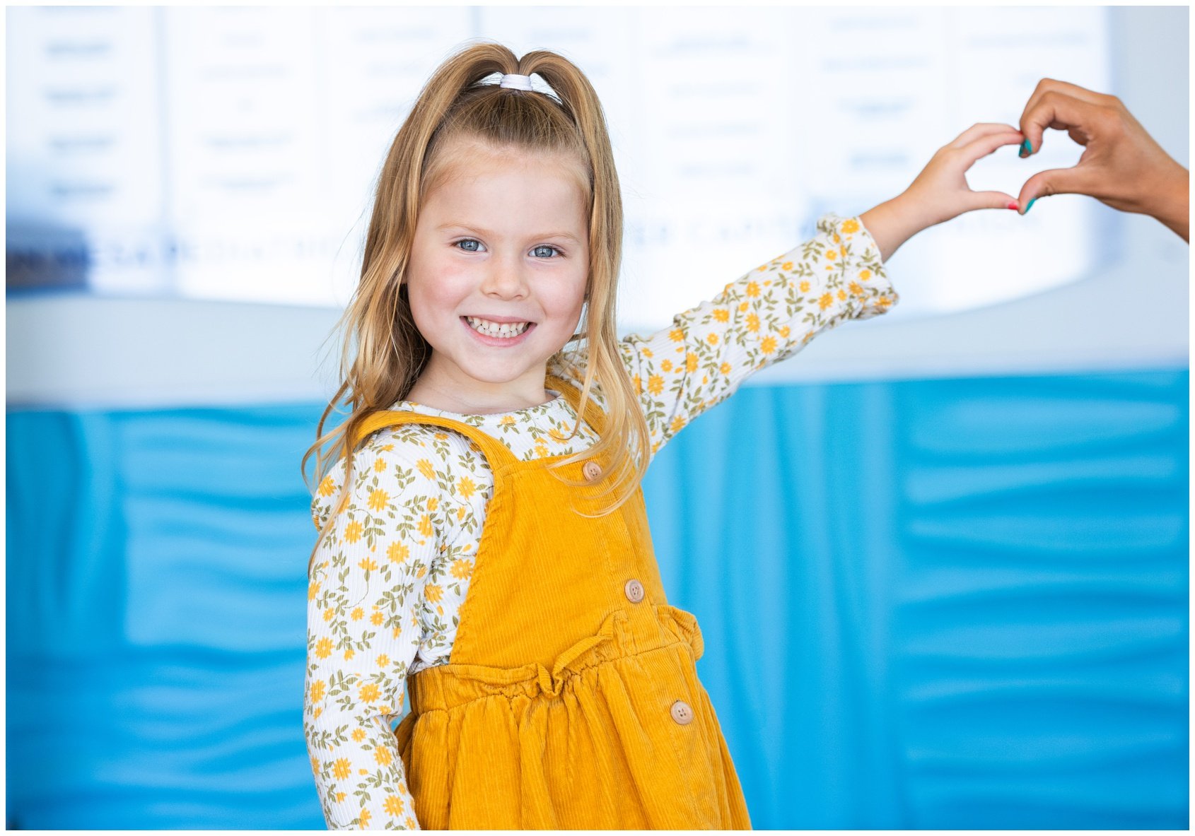 A smiling young girl with blonde hair in a ponytail, wearing a yellow pinafore dress over a floral shirt, making a heart shape with hands.