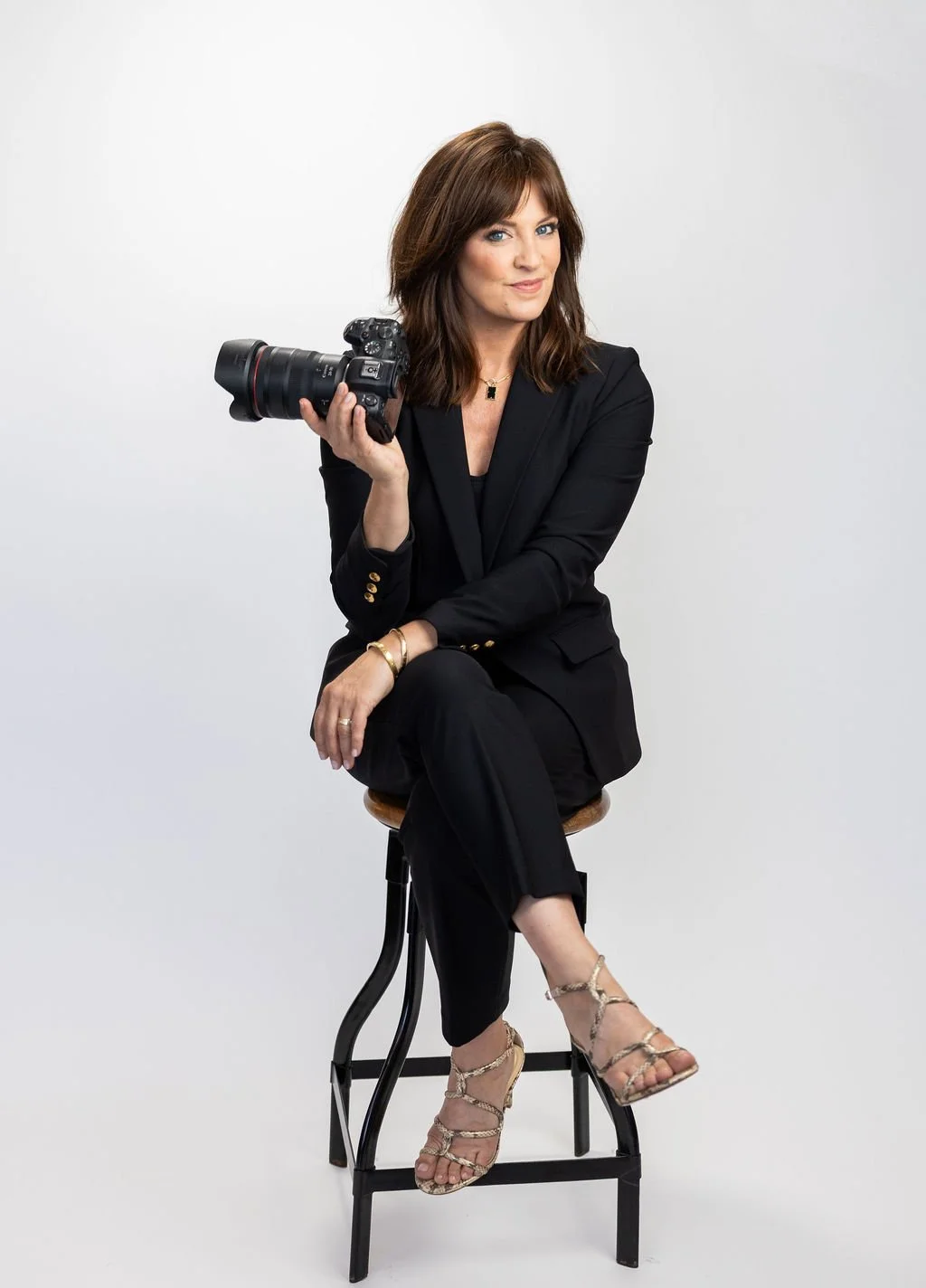 A woman with brunette hair wearing a black suit and beige high heel sandals, holding a camera, sitting on a stool against a plain white background.
