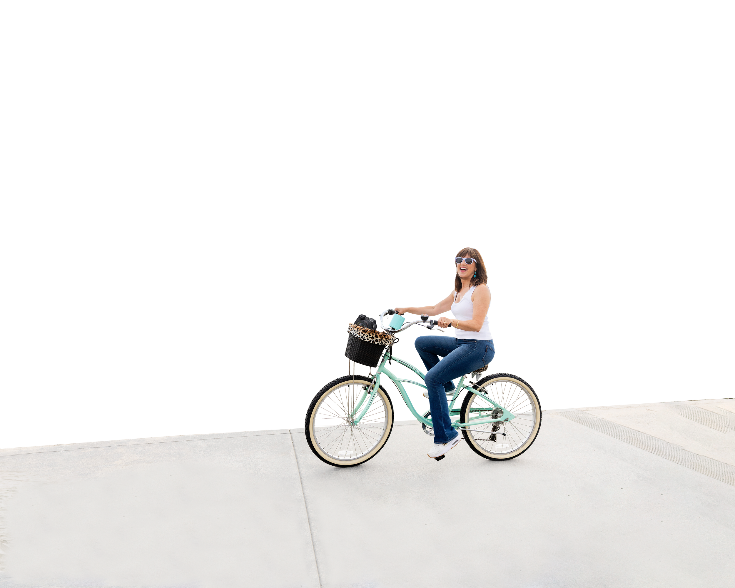 Young woman with brown hair wearing sunglasses, white tank top, and jeans riding a light green bicycle with a leopard print basket on a sidewalk.