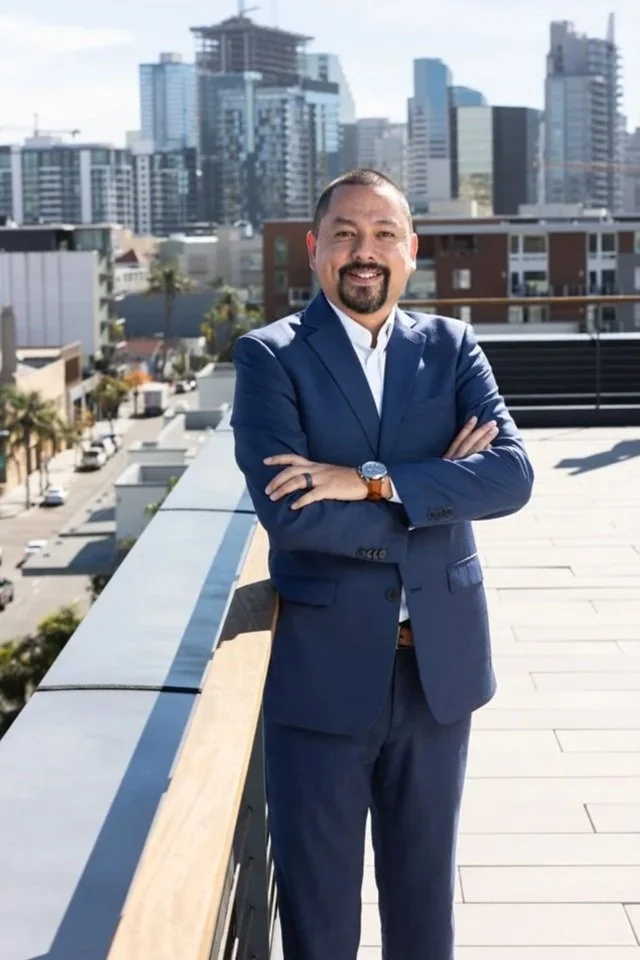 A man in a navy blue suit standing on a rooftop balcony with arms crossed, smiling, with a city skyline in the background.