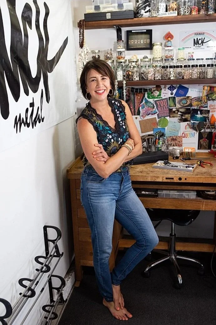 Smiling woman with short brown hair, wearing a sleeveless black top with colorful floral design, blue jeans, and bracelets, standing in a craft or art studio with shelves of jars and supplies behind her.