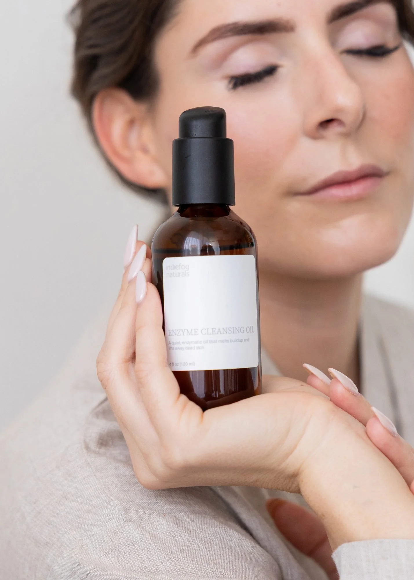 A woman with short brown hair and closed eyes holds a brown bottle labeled 'Enzyme Cleansing Oil' close to her face. She is sitting against a neutral background and appears relaxed.