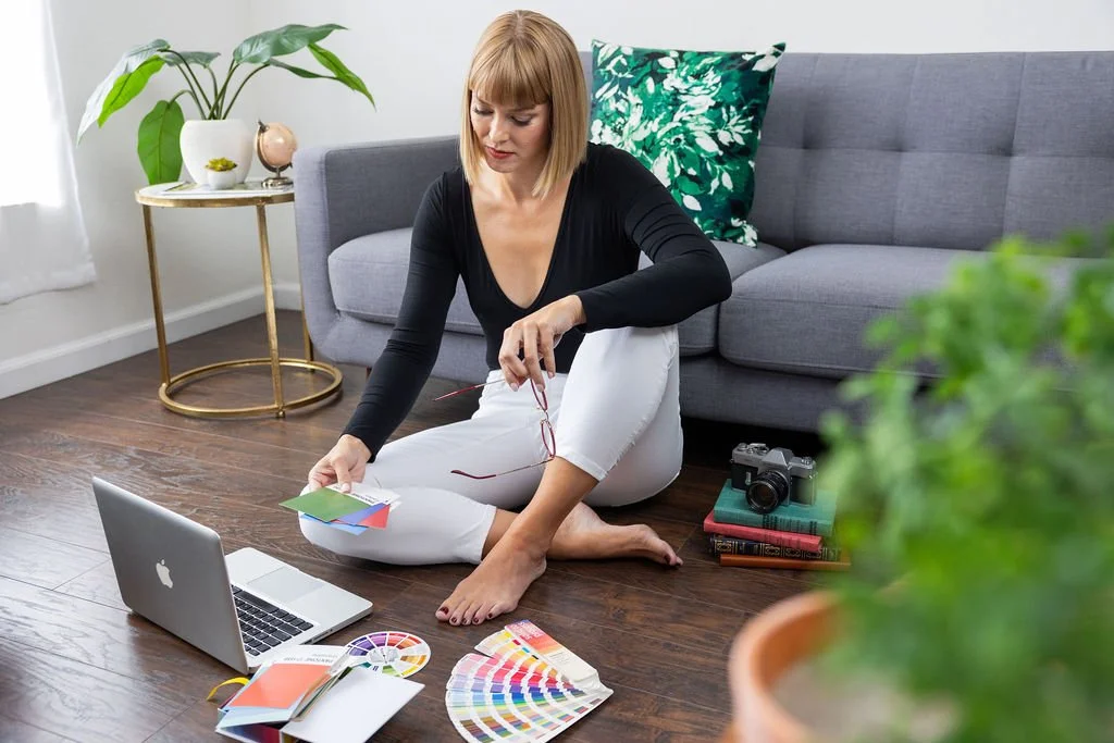 A woman sitting on the floor in a living room with a laptop, color swatches, and design tools, preparing for a project.