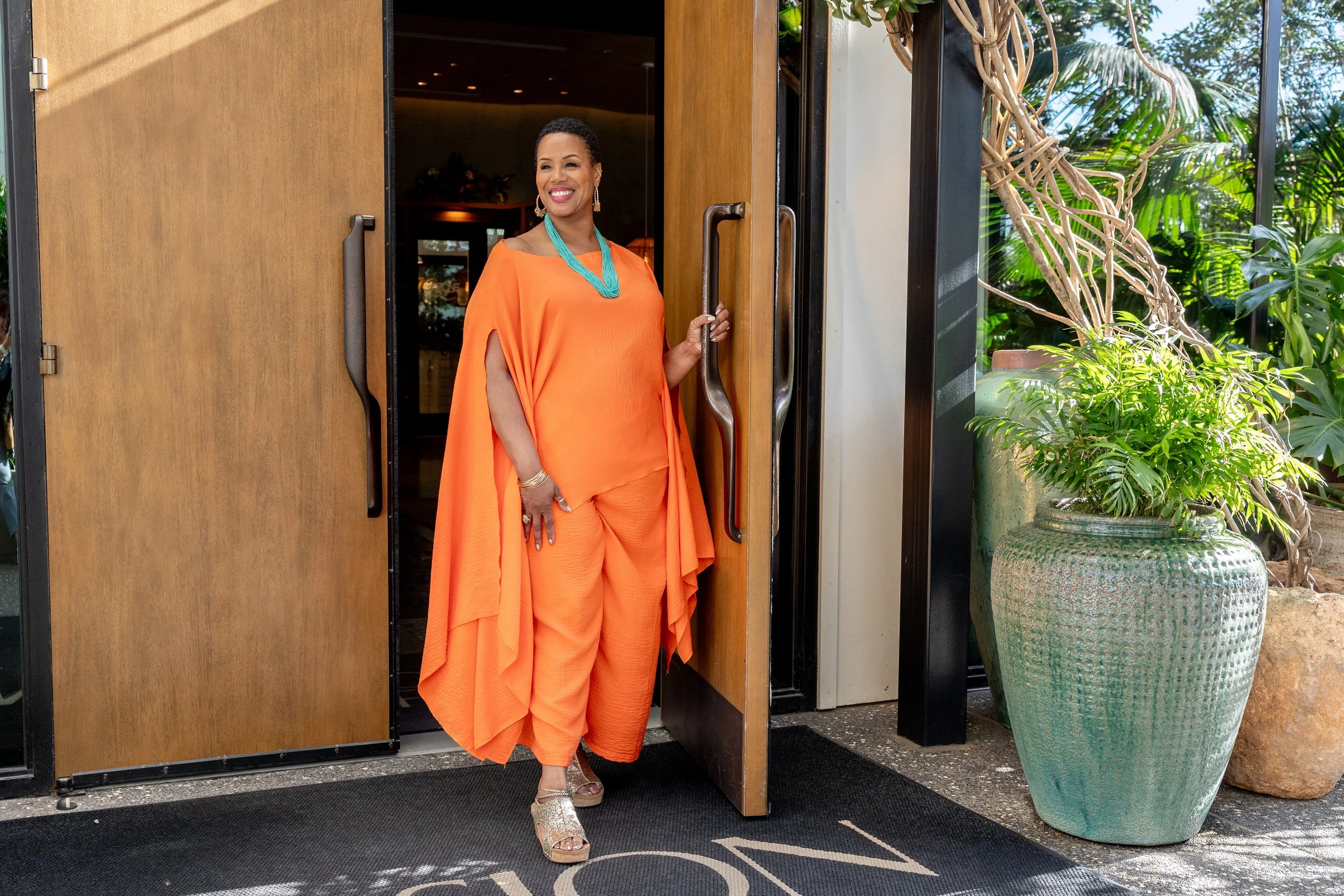A woman in bright orange, flowing clothing stands at an open doorway, smiling, with tropical plants in a large pot and lush greenery outside.