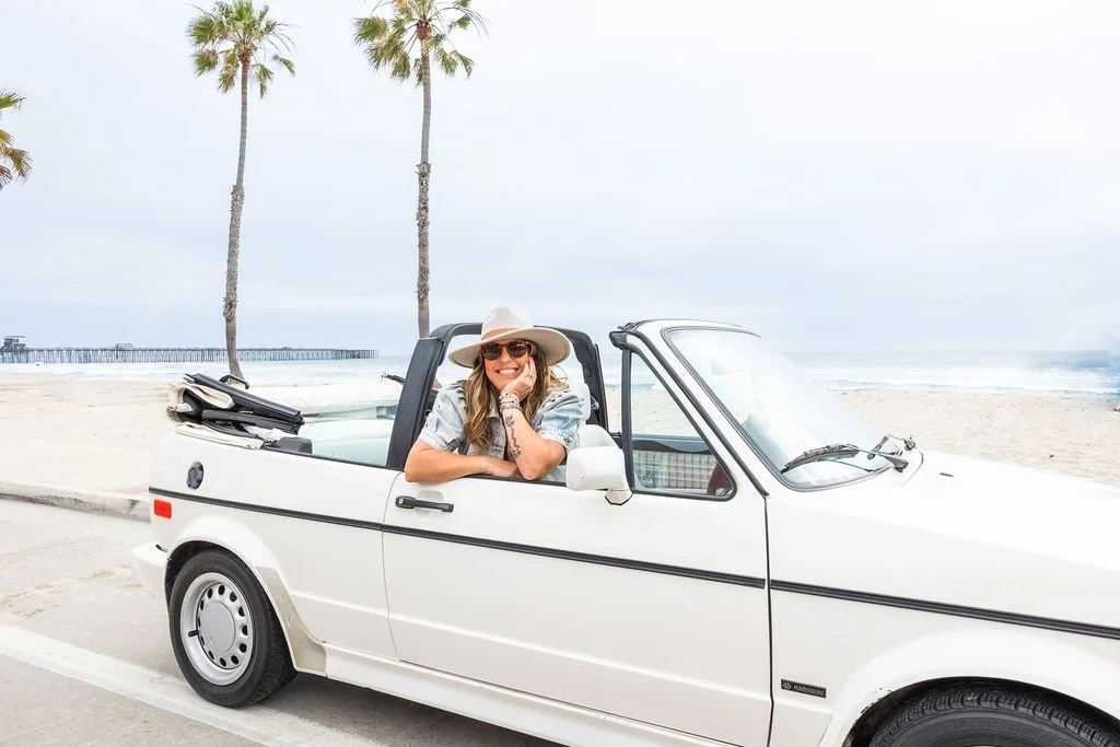 A woman with long hair, wearing sunglasses, a wide-brimmed hat, and a patterned shirt, is smiling and leaning out of the driver's side window of a white convertible car parked on a beachside road. There are palm trees and the ocean in the background.