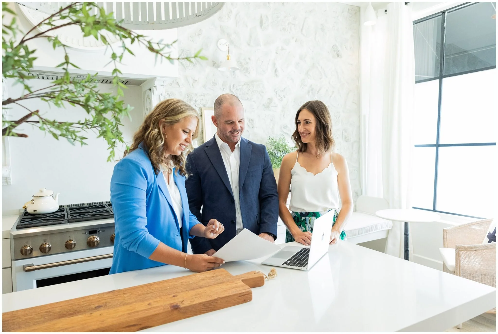 Three people, two women and a man, standing in a bright modern kitchen, looking at papers and smiling.