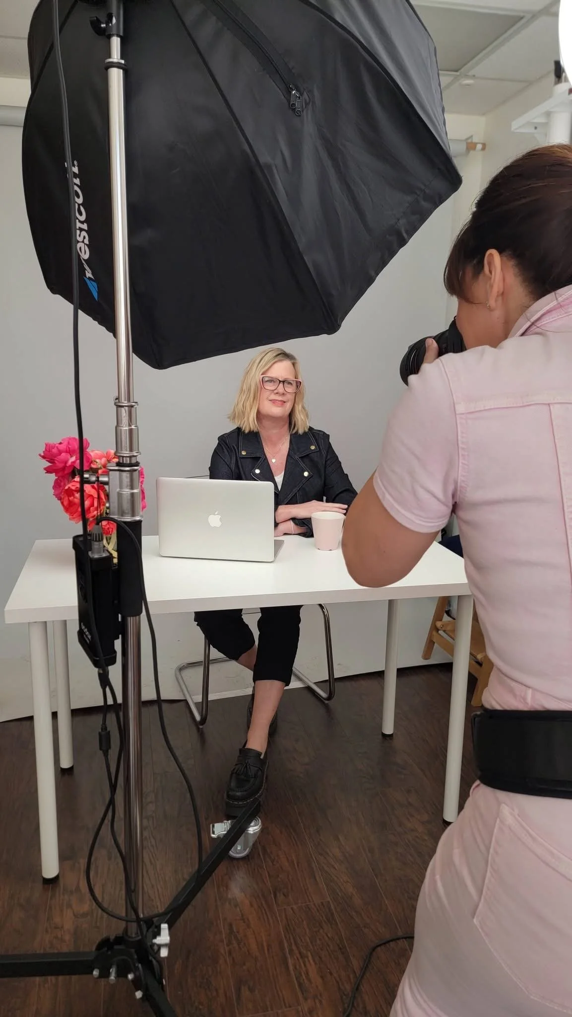 A woman with blonde hair and pink glasses sitting at a white table, being photographed by another woman with dark hair taking a picture with a camera. There is a softbox light overhead, a laptop, a pink mug, and a vase with pink flowers on the table.