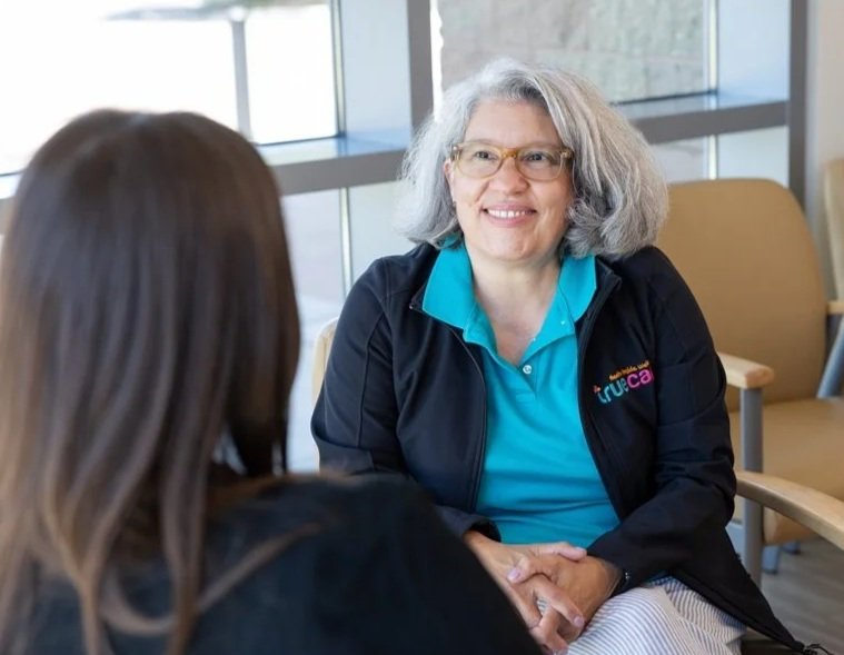Two women engaged in conversation in a room with large windows; one woman is smiling, wearing glasses, and a black jacket with a colorful logo, while the other has long brown hair and is facing away from the camera.