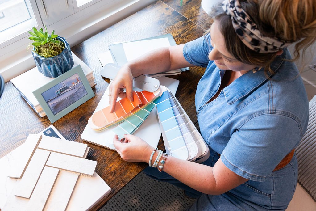 A woman at a desk reviewing a color palette for interior design or home renovation, with paint swatches and color samples, near a window with a potted plant and framed photo.