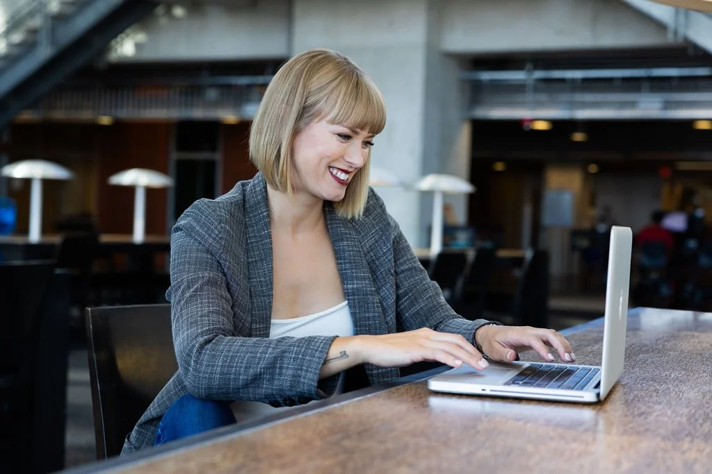 A woman with shoulder-length blonde hair, wearing a gray plaid blazer and white top, sits at a table in a public space, smiling while using her laptop.