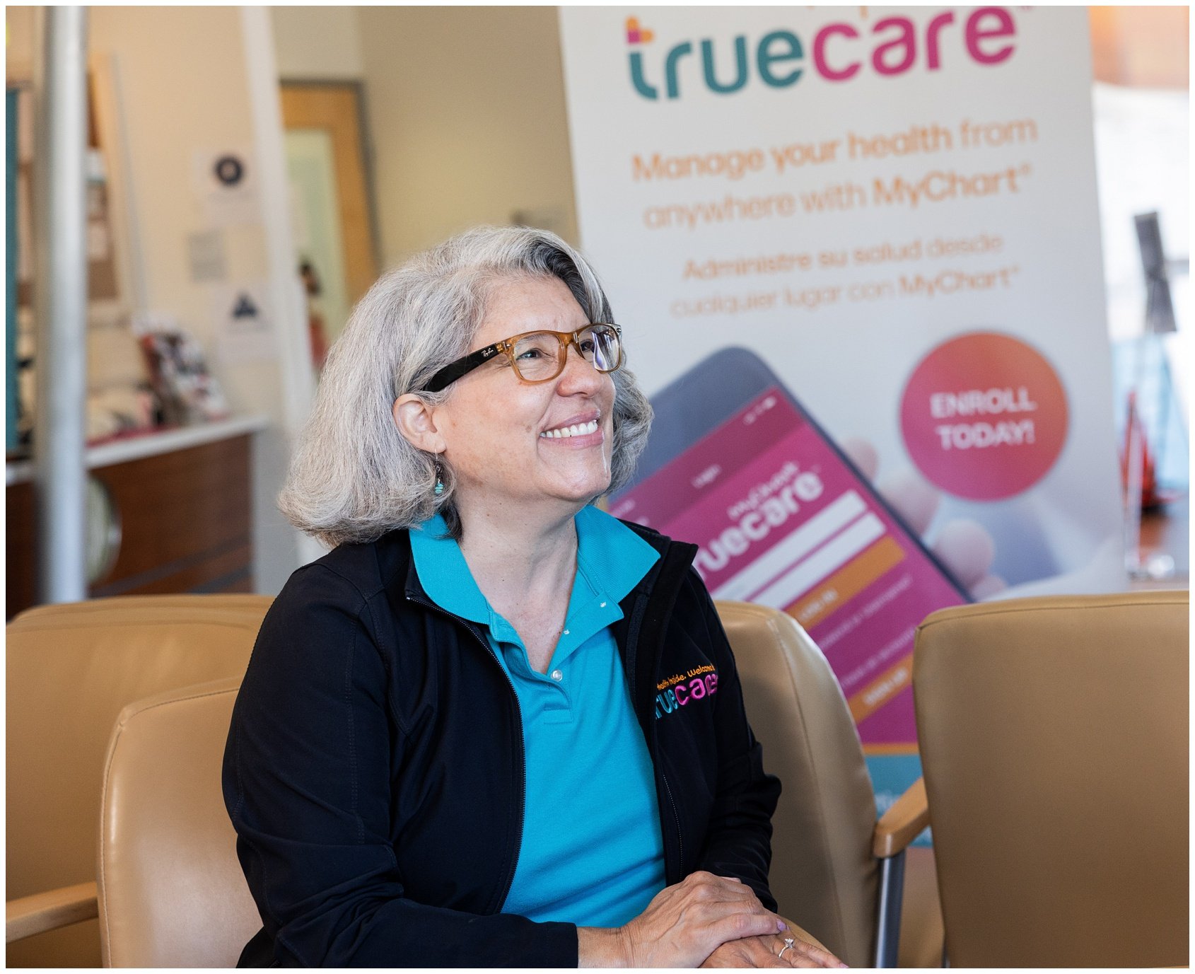 A smiling woman with gray hair, glasses, and a blue collared shirt, sitting in a waiting area. Behind her is a health care promotional poster with the words "truecare" and a mobile app image.