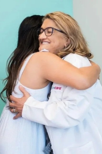 Two women hugging and smiling in a friendly embrace, one wearing a white dress and the other in a medical coat.