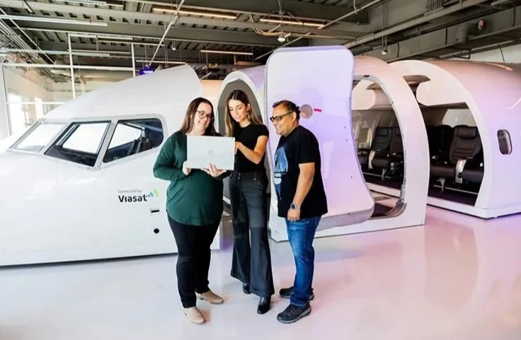 Three people standing next to a flight simulator shaped like an airplane cockpit, looking at a laptop.