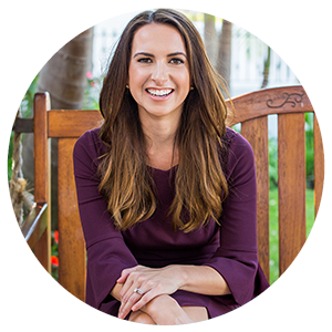A woman with long brown hair wearing a purple top, smiling and sitting outdoors on a wooden chair.