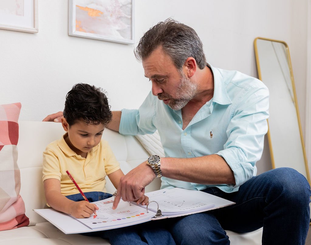 A man and a young boy sitting on a white couch, looking at a workbook or folder together. The man is pointing at the workbook, and the boy is holding a red pencil, appearing focused. The background includes framed artwork and a yellow-framed mirror.