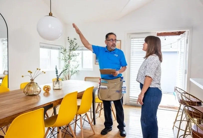 A man in a blue shirt and beige apron talks to a woman with brown hair wearing a white polka dot blouse inside a bright, modern dining area with yellow chairs and decorative vases on the table.