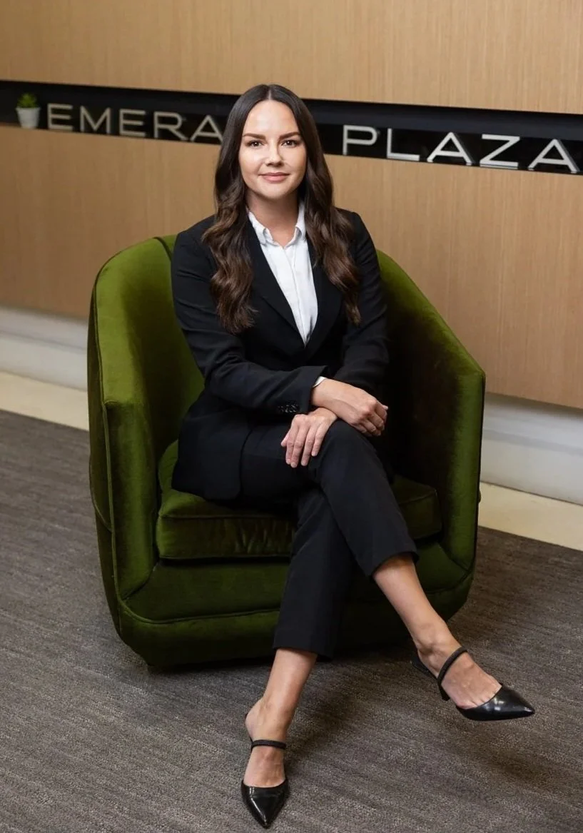 A woman sitting on a green velvet armchair, wearing a black suit and white shirt, with long brunette hair, in a professional setting with a wooden wall background, and a sign that reads 'Emera Plaza'.