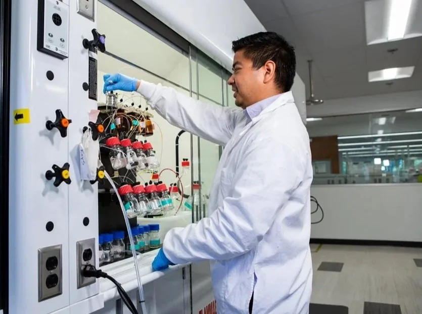 A scientist working in a laboratory, handling laboratory equipment behind a fume hood with various bottles and wires.