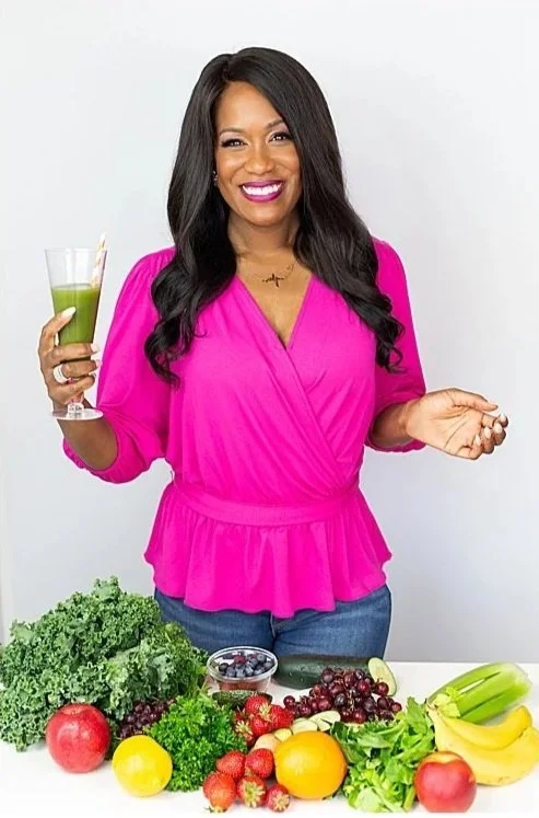 Smiling woman in a bright pink top holding a glass of green juice, standing behind a table with various fresh fruits and vegetables.