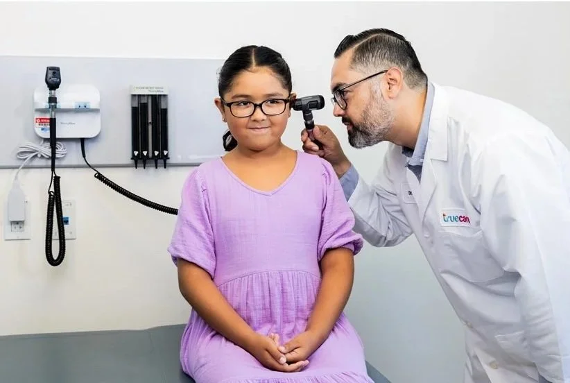 A young girl sitting on an examination table at a doctor's office, with a doctor examining her ear using an otoscope.