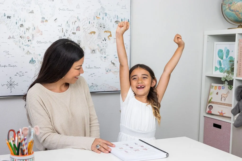 A young girl and woman sitting at a desk, smiling, with the girl raising her arms in excitement. The girl is wearing a white dress, and the woman has long dark hair and is wearing a beige sweater. There are art supplies in a cup on the desk, and a world globe and books on a shelf in the background.