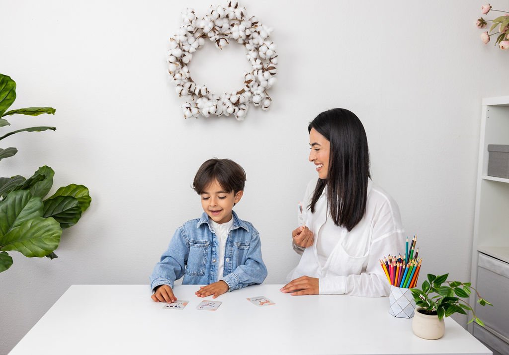 A woman and a young boy sitting at a white table playing a card game indoors. The woman has dark hair and wears a white shirt, smiling at the boy. The boy has brown hair, wears a denim jacket, and is focused on the cards. There are green plants and a cotton wreath decoration on the wall behind them.
