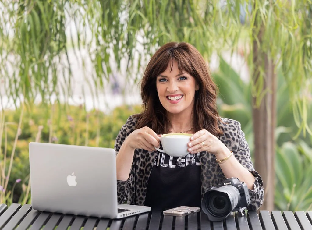 A woman with brown hair and a leopard print blazer sitting outdoors at a table with a laptop and camera, holding a coffee mug and smiling.