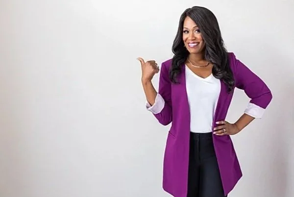 A woman with dark hair wearing a purple blazer, white shirt, and black pants, smiling and pointing at herself against a plain white background.