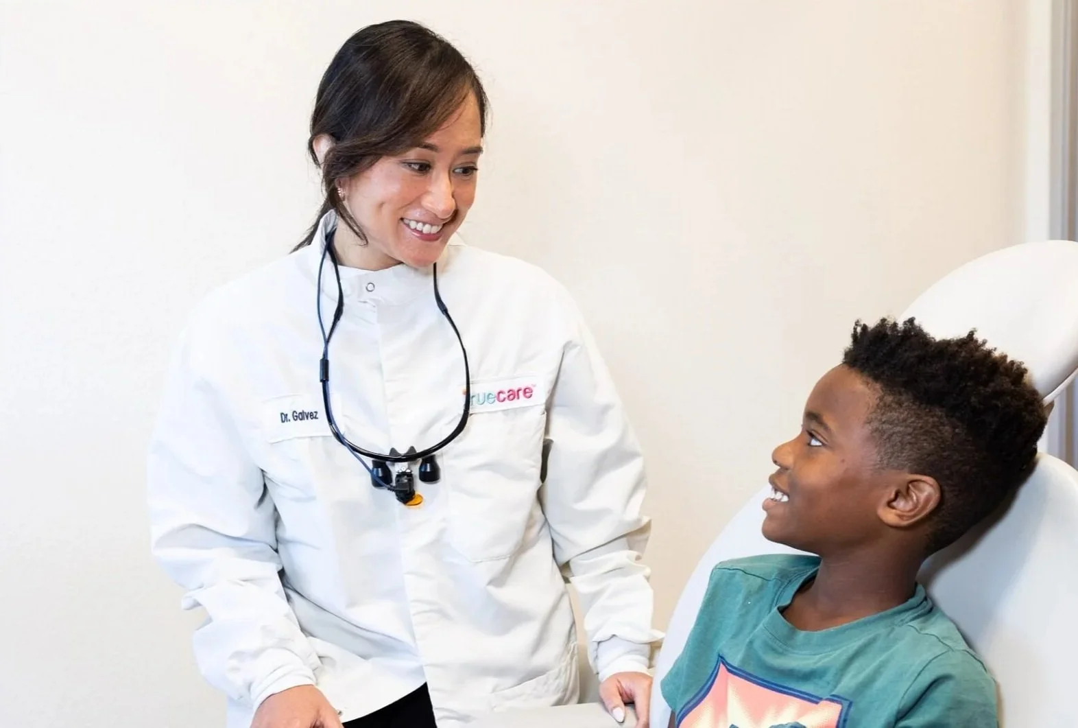 A female healthcare professional in a white coat smiling and talking to a young boy sitting in a hospital bed, wearing a teal shirt, with a cheerful expression.