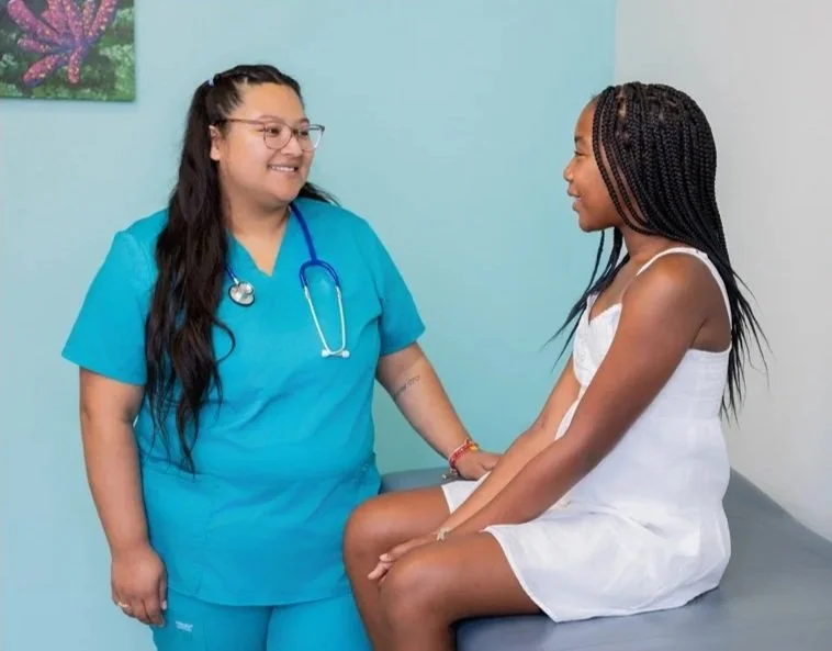 A healthcare professional, wearing blue scrubs and a stethoscope, smiling at a young girl in a white hospital gown who is sitting on an examination table and holding the nurse's hand.