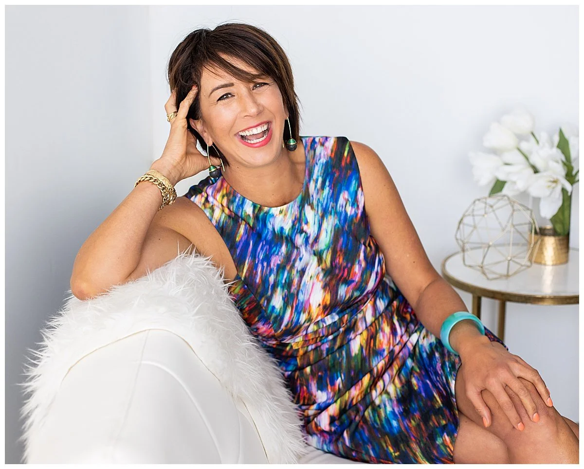 A woman with short brown hair sitting on a white furry chair, smiling and laughing, wearing a colorful sleeveless dress, gold jewelry, and a turquoise bracelet. There is a side table with white flowers and geometric decor behind her.