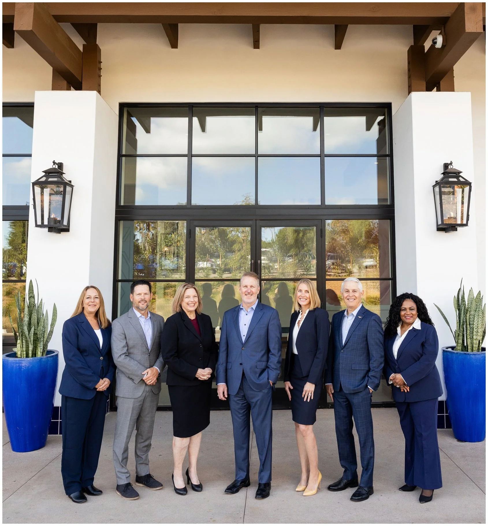 A group of eight diverse professionals standing outside in front of a modern glass building, smiling for a photo. The group includes both men and women dressed in business attire, with two women at either end wearing dark blue blazers and the others 