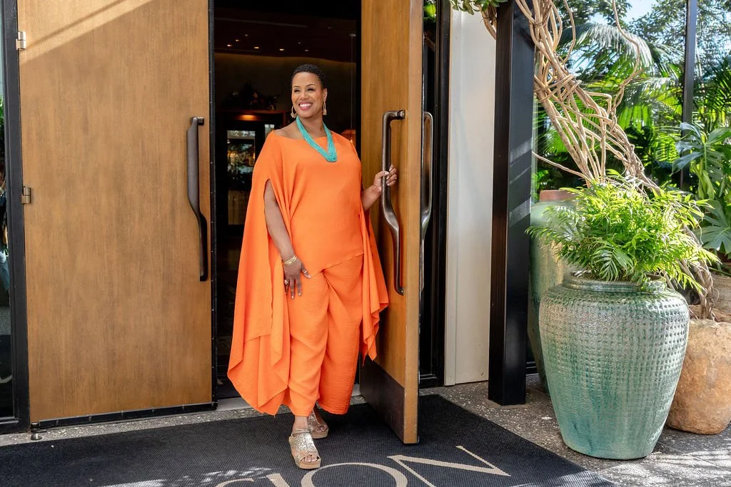 A woman in an orange outfit standing at an open door, smiling. She is wearing jewelry and sandals, with lush green plants in a large pot nearby.