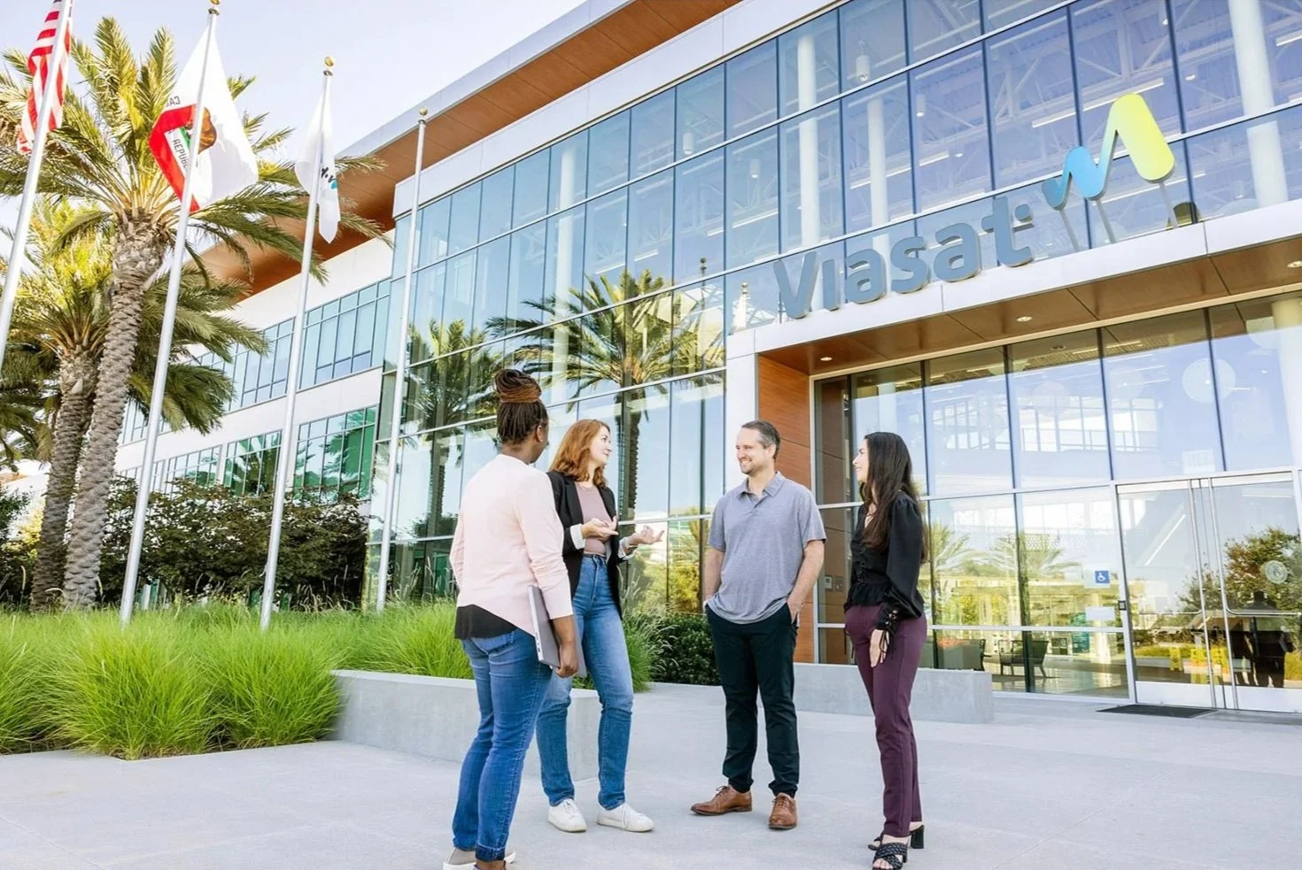Four people standing and talking outside the Masat building, a modern glass office building with palm trees nearby and flags, in sunny weather.