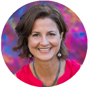 A smiling woman with short brown hair, wearing a pink top and a beaded necklace, in front of a colorful, blurred background.