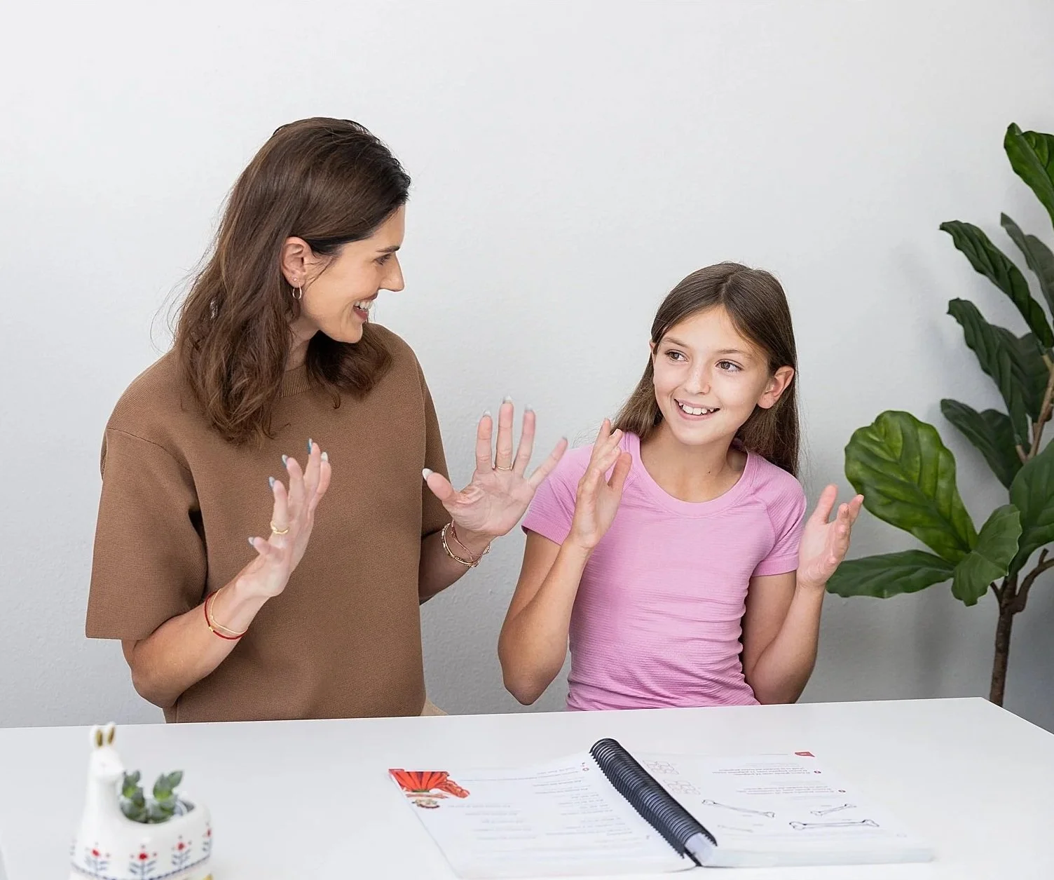 A woman and a girl smiling and high-fiving each other at a white table with papers, a pen, and a small potted plant, in a bright room with a green plant in the background.
