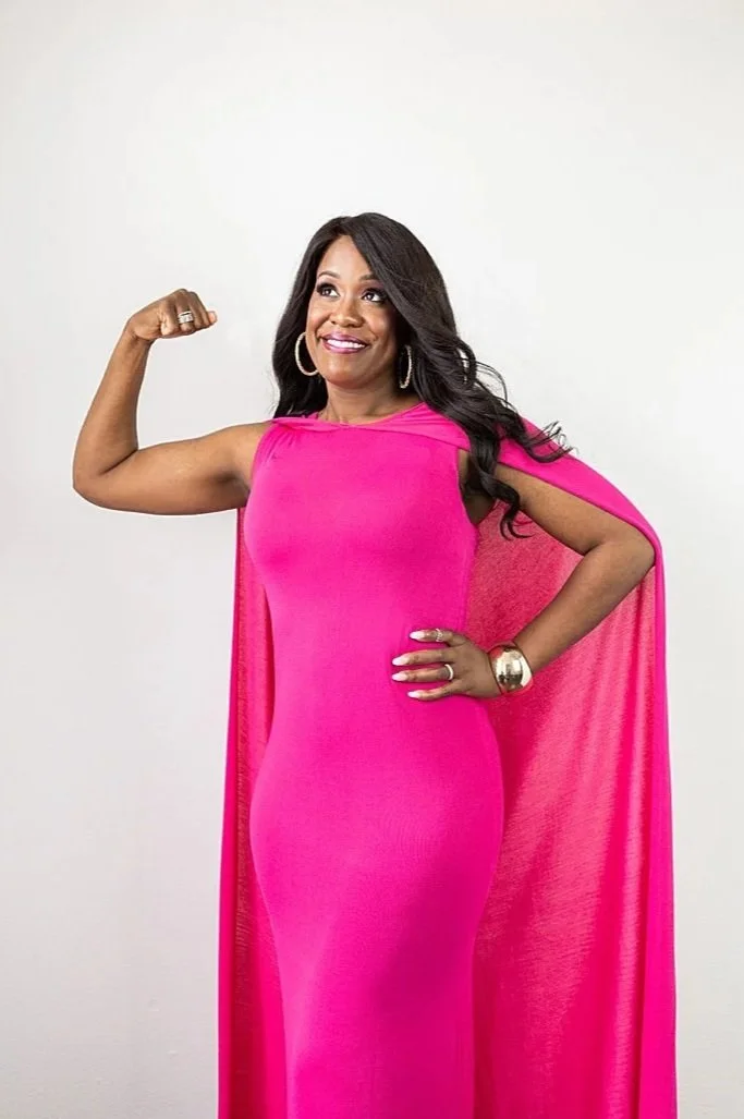 A woman wearing a bright pink, sleeveless, floor-length dress with a cape, smiling and flexing her right arm.