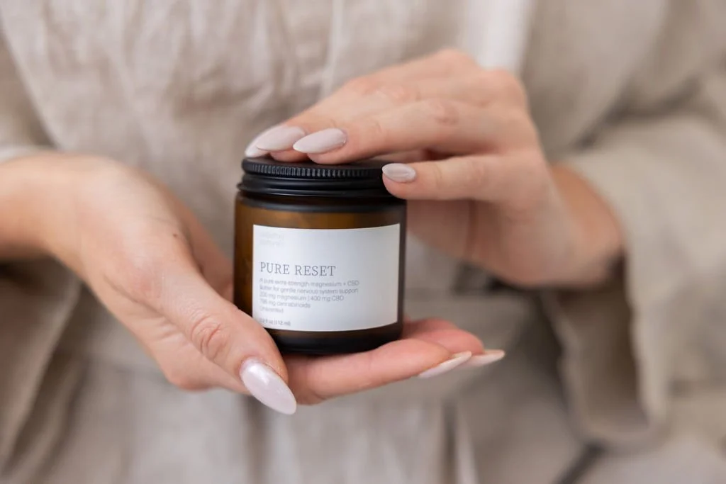 Person holding a small brown jar labeled 'PURE RESET' with a white label, in front of a neutral fabric background.