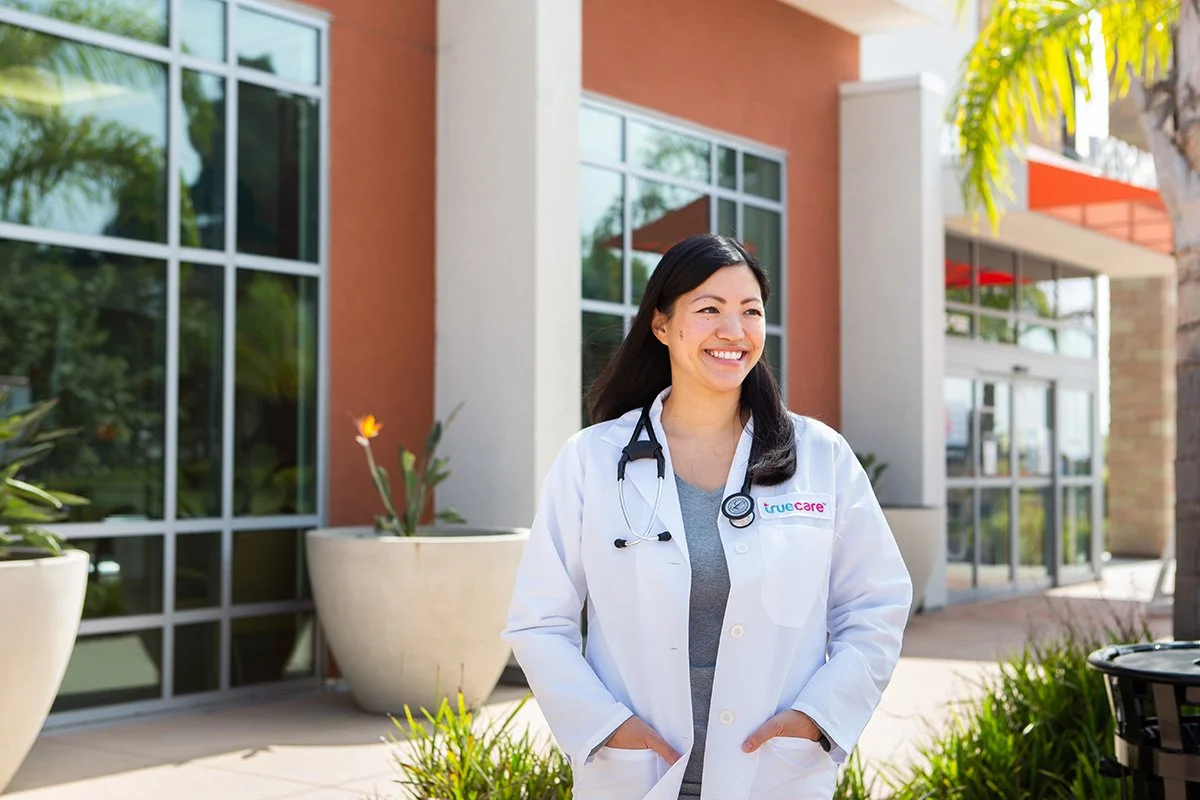 A smiling female doctor outdoors in front of a medical building, wearing a white coat with a stethoscope around her neck.