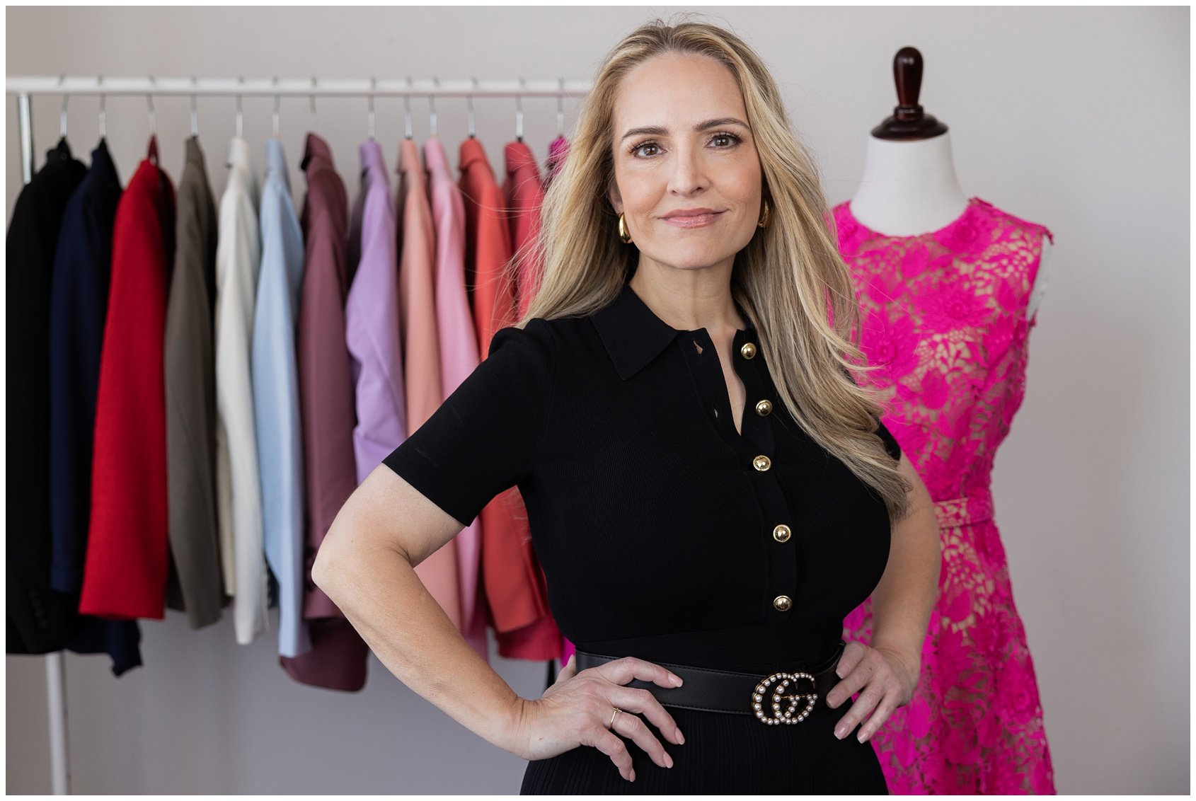 A woman with blonde hair and a black dress with gold buttons standing in front of a rack of colorful shirts and a pink lace dress on a mannequin.