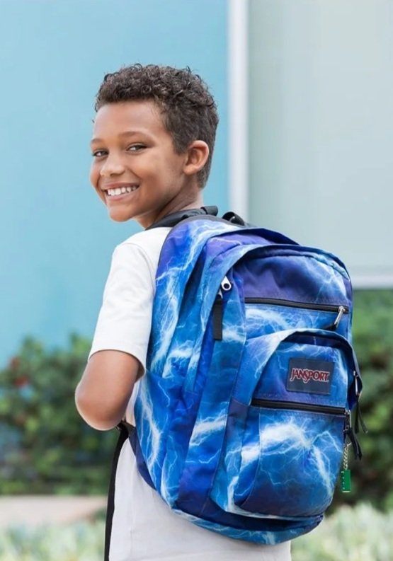 A young boy with curly hair smiling while wearing a white shirt and carrying a blue backpack with white streaks, standing outdoors.
