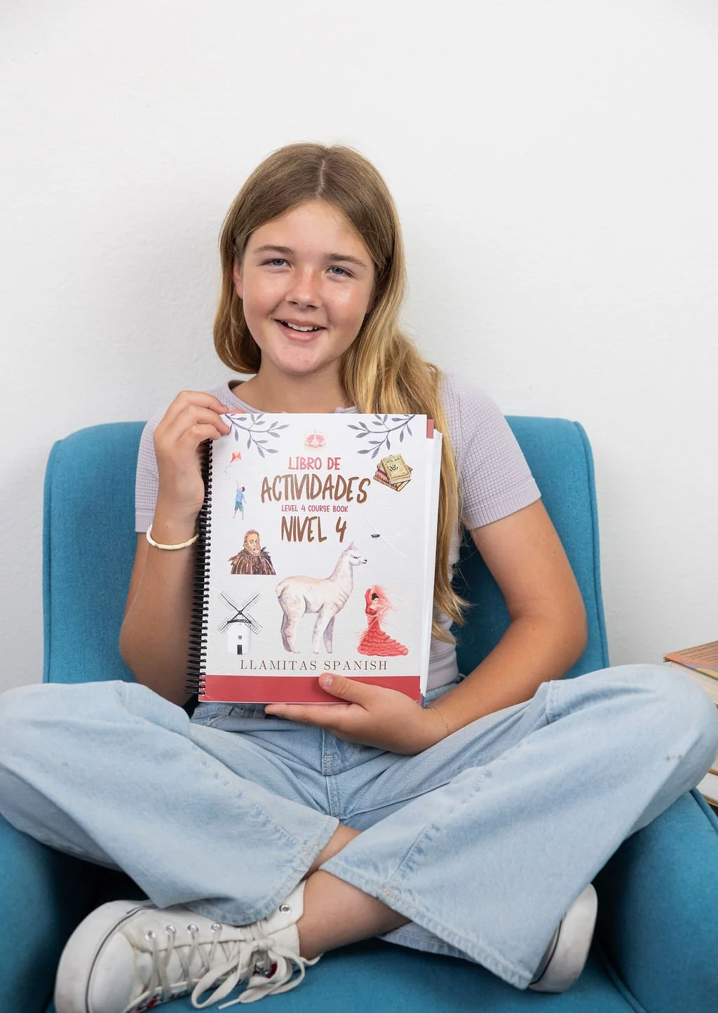 A young girl with long blonde hair wearing a gray t-shirt, light blue jeans, and white sneakers, sitting cross-legged on a blue chair, holding a Spanish activity book titled 'Libro de Actividades Nivel 4'.