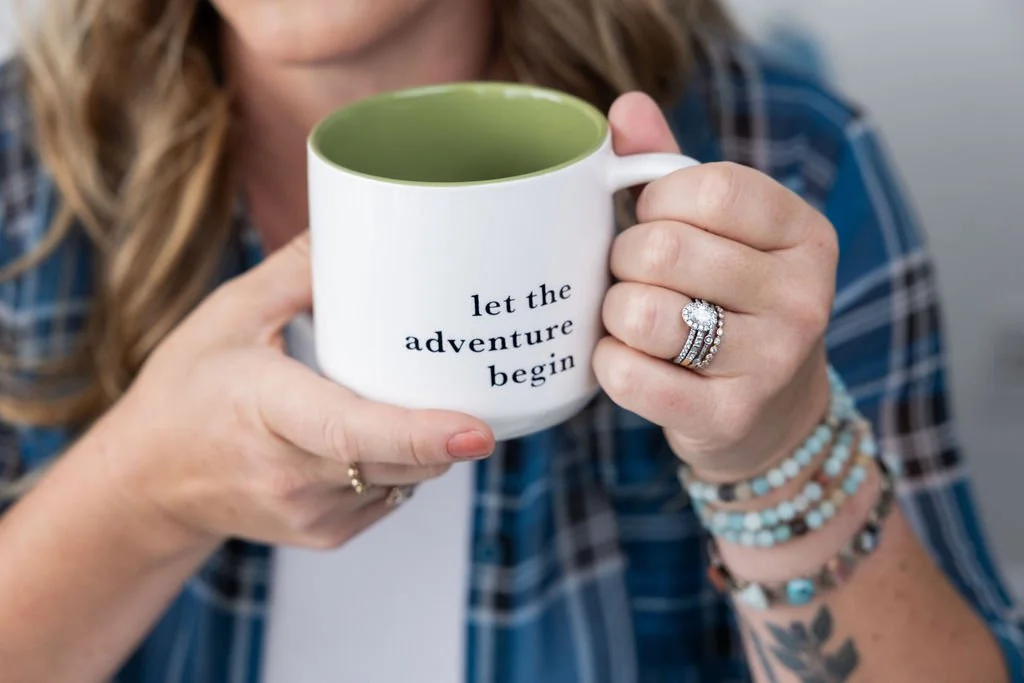 Woman holding a white mug with green interior and the message "let the adventure begin" printed on it, wearing rings and bracelets.